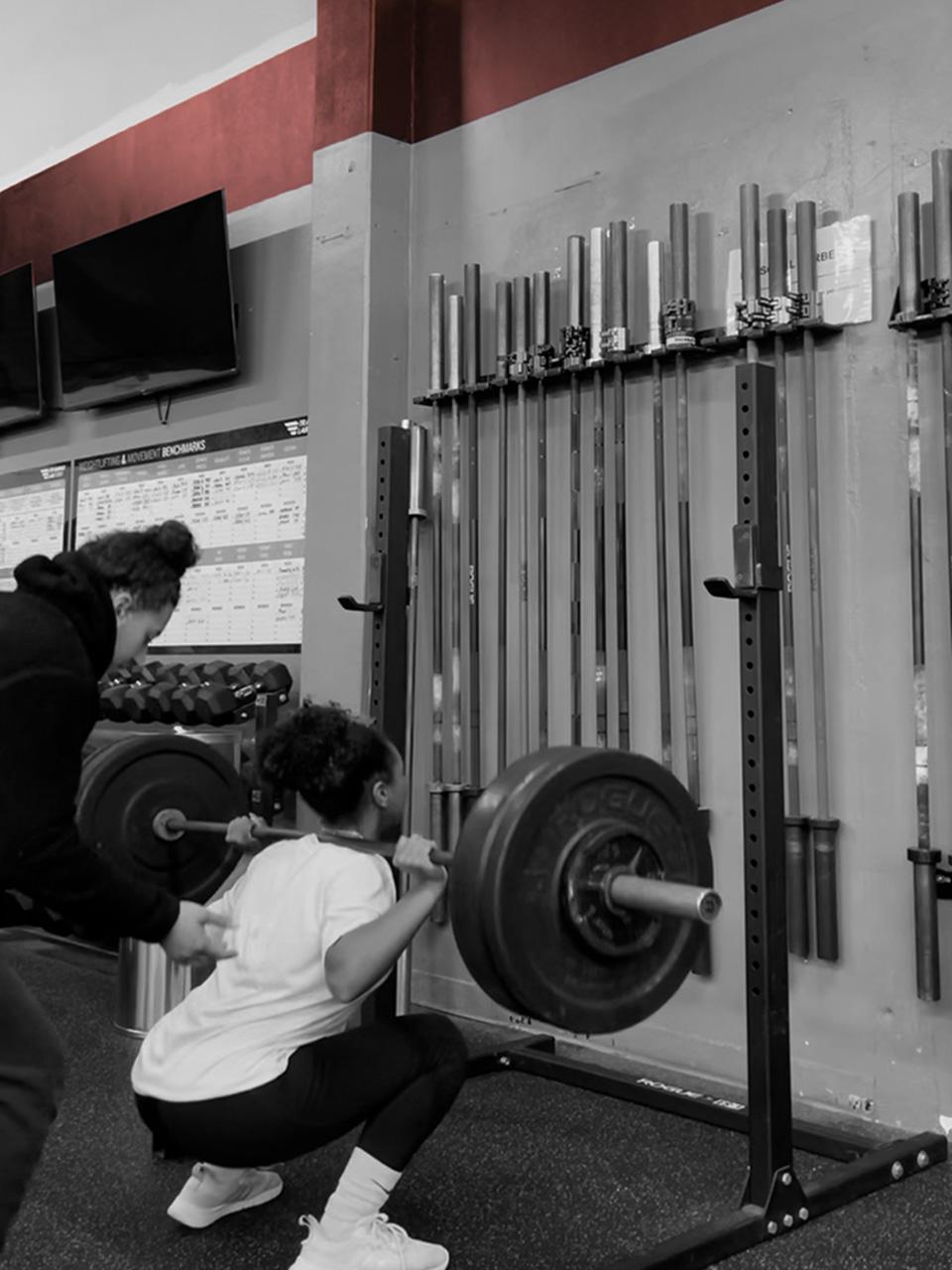 Athlete performing a barbell squat with a coach's support in a gym.